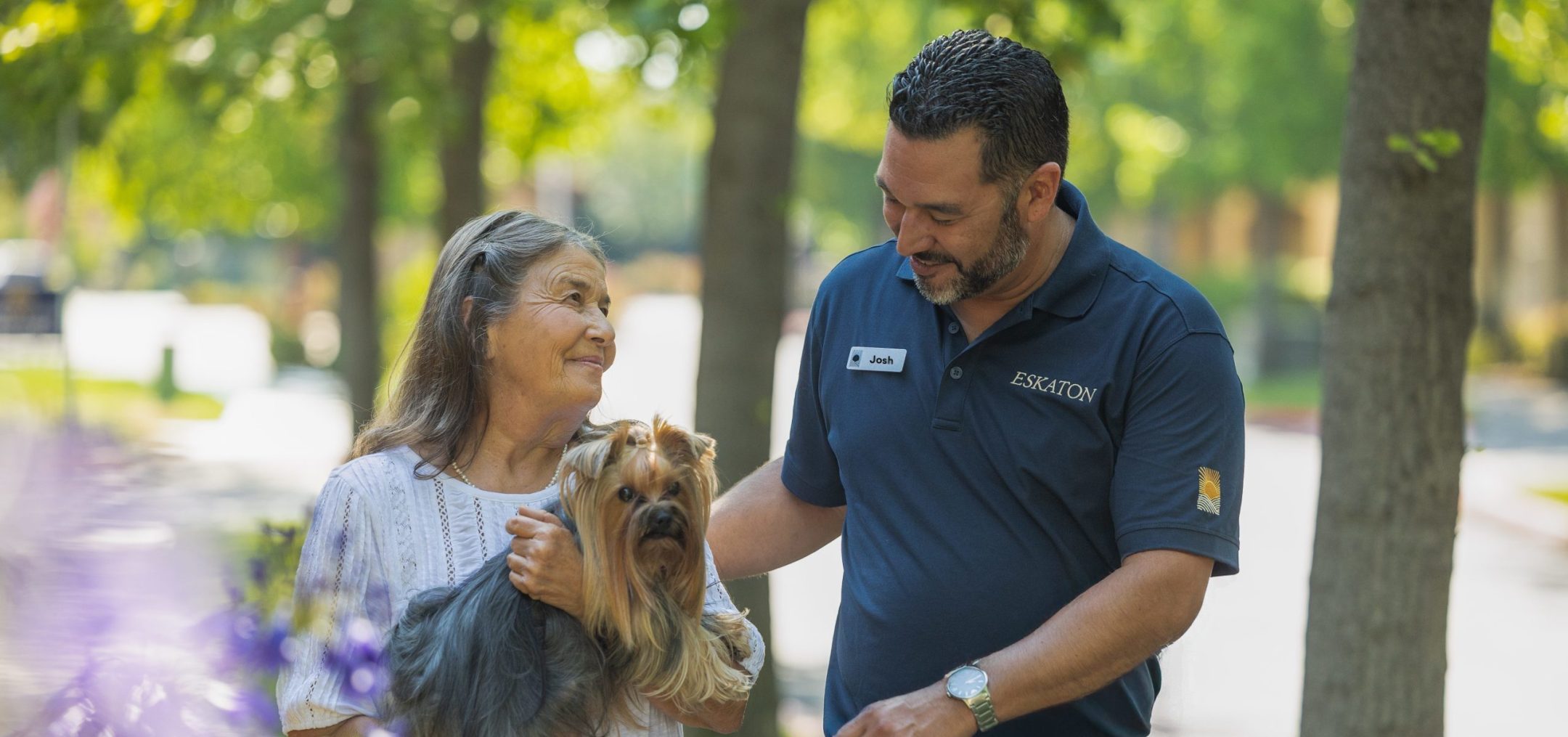An elderly woman holding a small dog walks outside with a smiling man in a navy polo shirt. They appear to be enjoying a pleasant conversation on a sunny, tree-lined path.