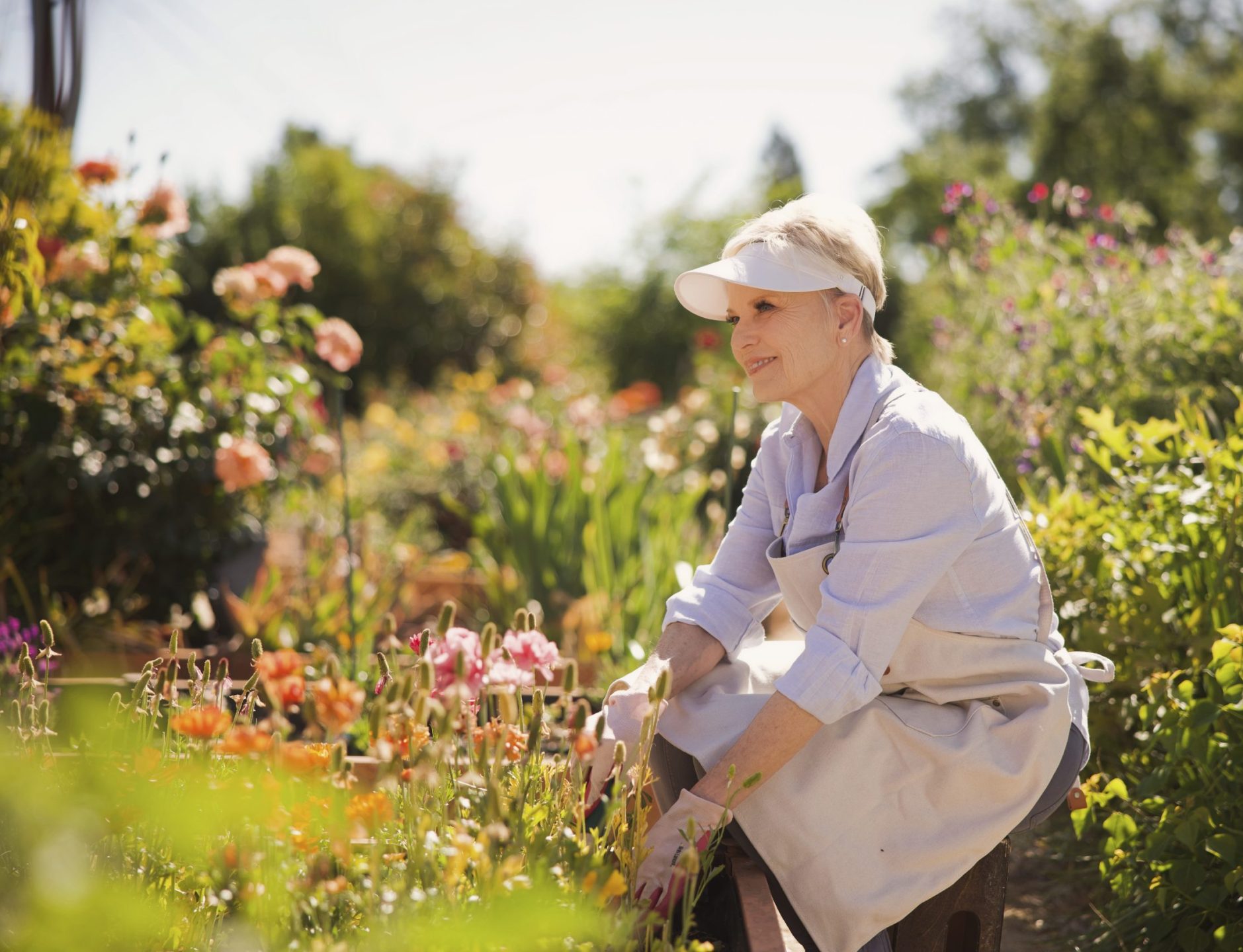 A woman wearing a sun visor and apron kneels in a bright, colorful garden, tending to flowers on a sunny day. Immersed in the resident culture of gardening, she appears content and focused among blooming plants and greenery.