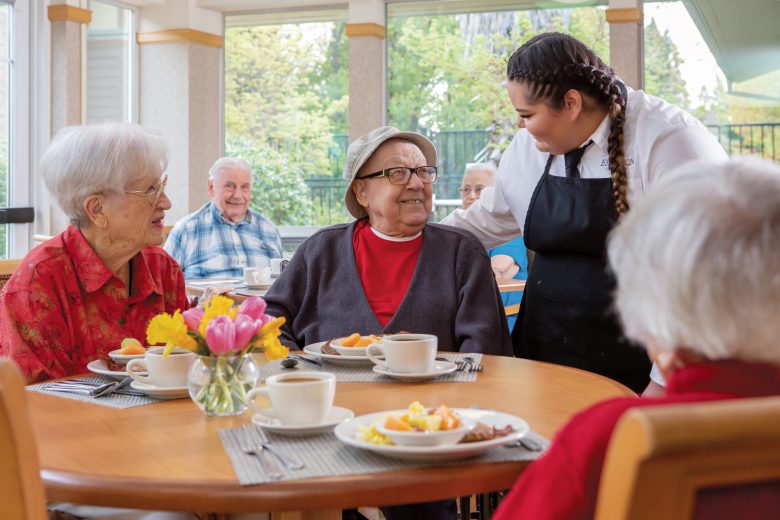 A smiling caregiver stands beside a table where three elderly people are seated, chatting and enjoying a meal together in a bright, cheerful dining room with large windows and flowers on the table.