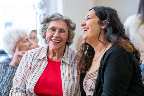 An elderly woman and a younger woman sit close together, smiling and laughing. Both appear happy and engaged, sharing a joyful moment indoors. Other people are visible in the softly focused background.