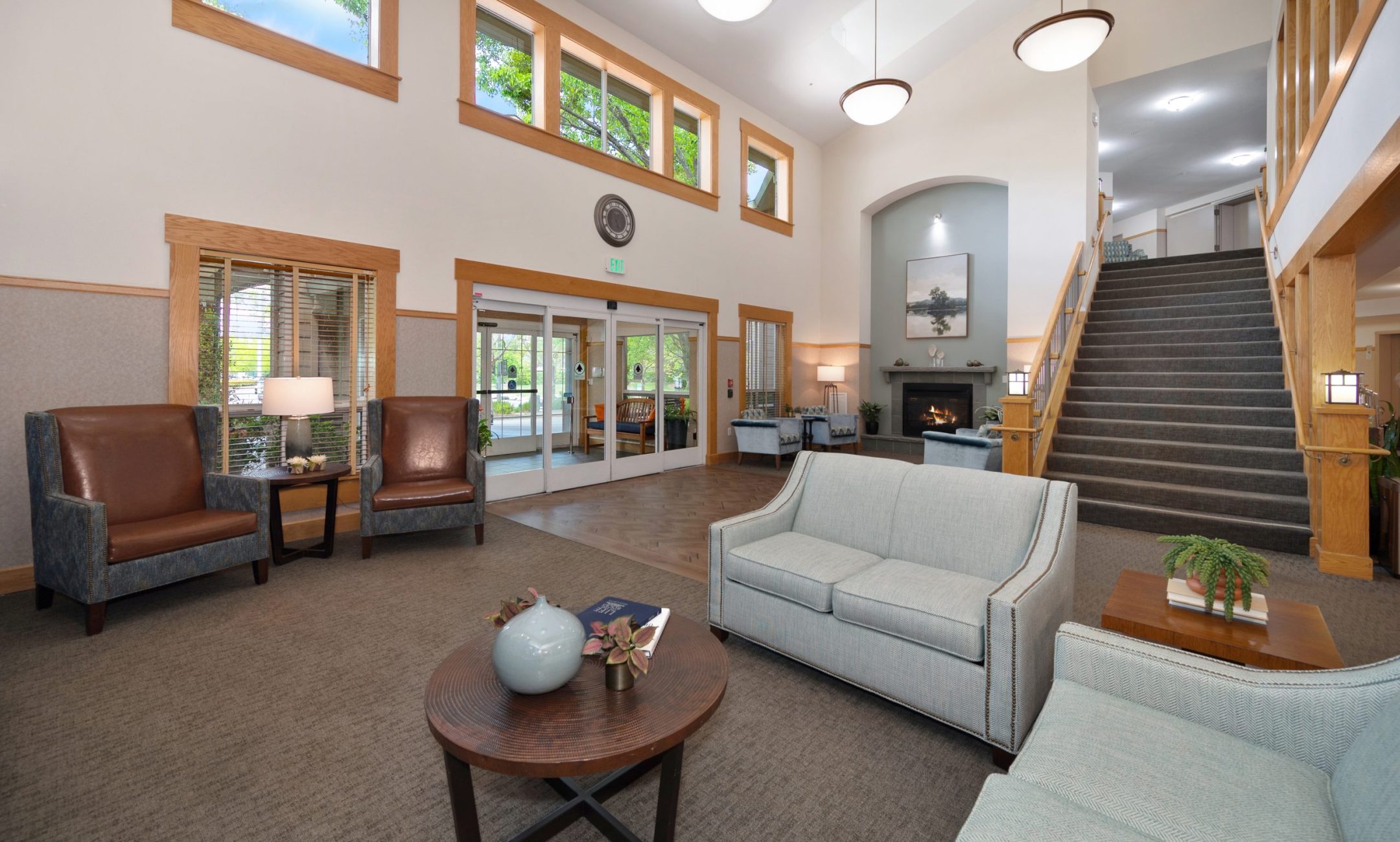 A spacious, well-lit lobby features comfortable seating, large windows, a staircase, and double glass doors. Modern light fixtures hang from the high ceiling, and a fireplace warms the back wall.