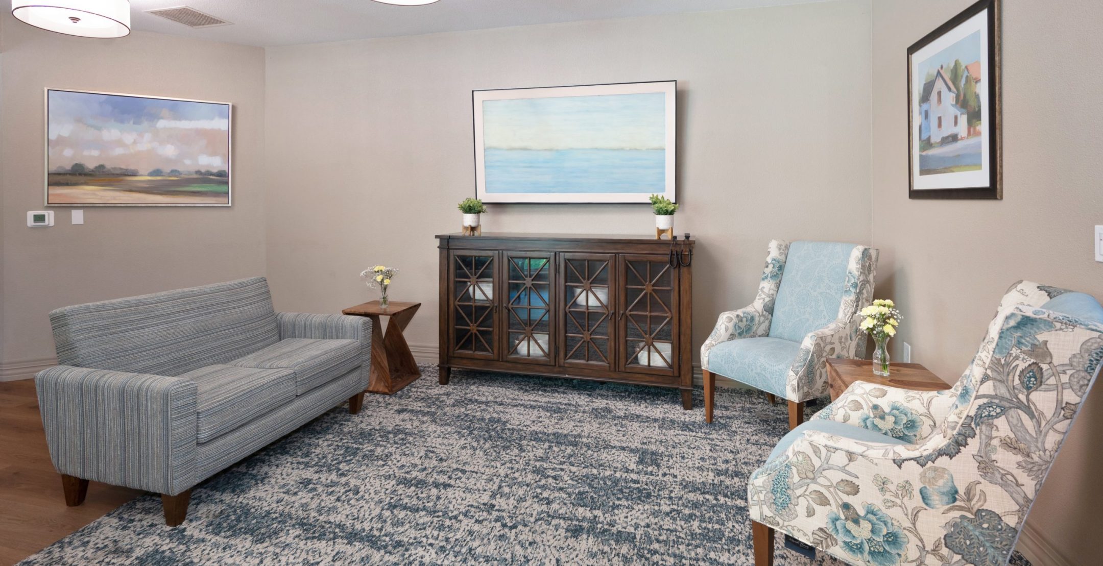 A cozy living room with a blue patterned rug, a gray loveseat, two upholstered armchairs, a wooden cabinet, small side table with flowers, and landscape artwork on light-colored walls under modern ceiling lights.