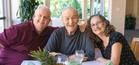 Three smiling adults sit closely together at a dining table, posing for a photo. The man in the center has gray hair, and they enjoy the Eskaton Gold River lifestyle indoors with green plants and sunlight shining through the windows behind them.