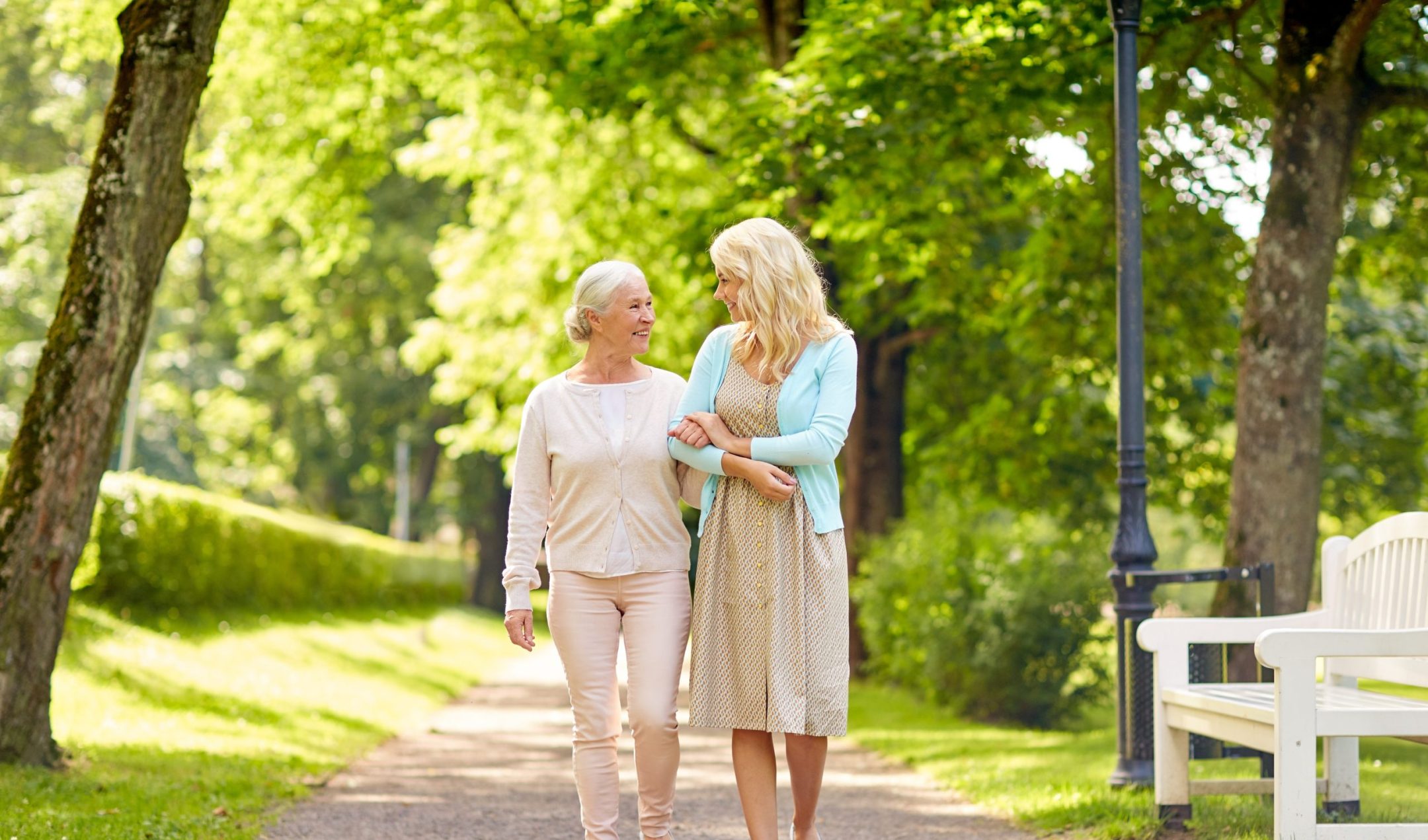 Two women, one older and one younger, walk arm-in-arm on a sunny park path lined with trees and greenery. Both are smiling and engaged in conversation, enjoying quality time together during this Pre-Memory Care stage.