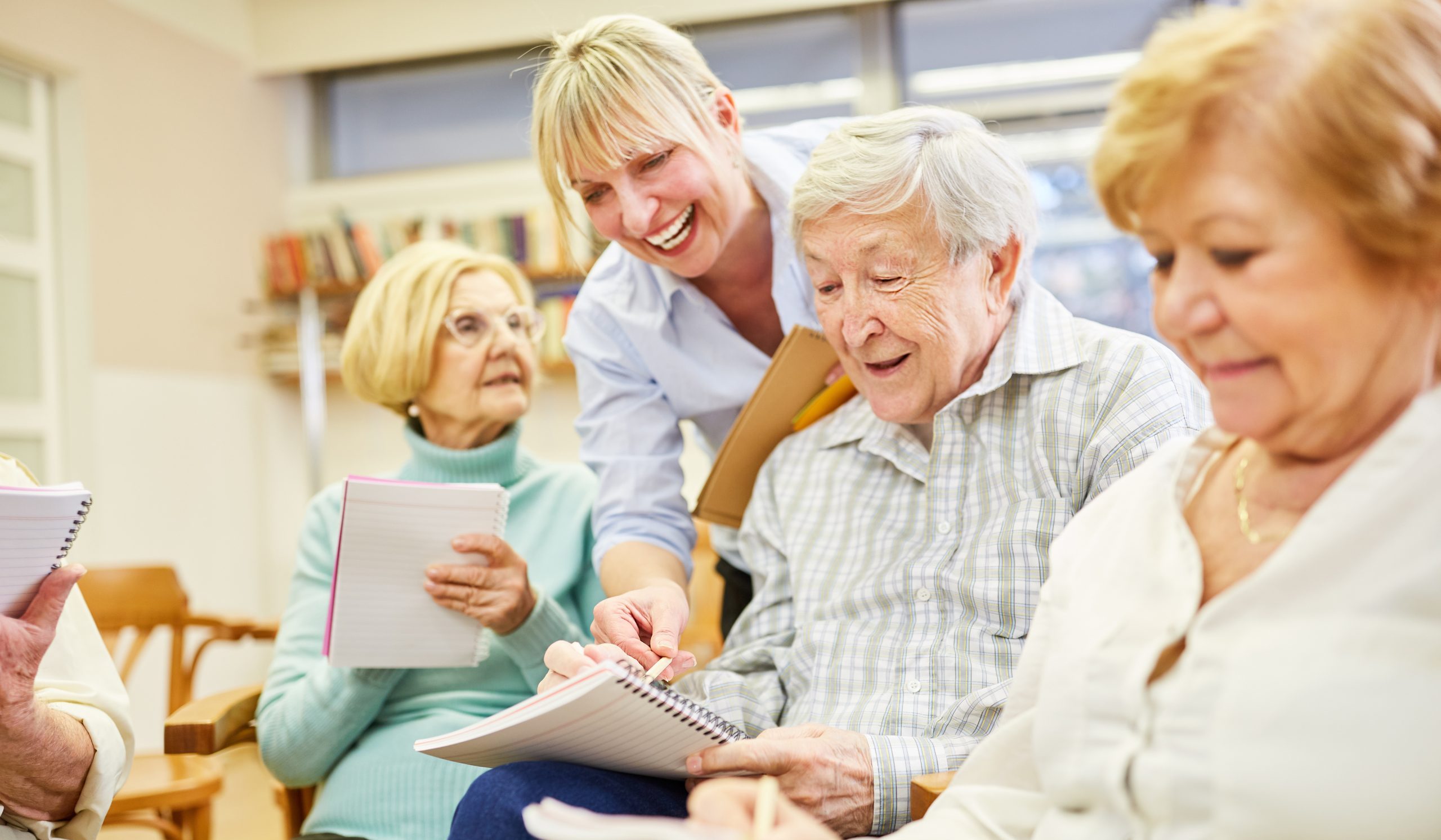 A group of smiling seniors sits together in a classroom at Eskaton Gold River memory care, holding notebooks and pens, while a woman stands by, assisting one of the men with his notes. The atmosphere is friendly and collaborative.