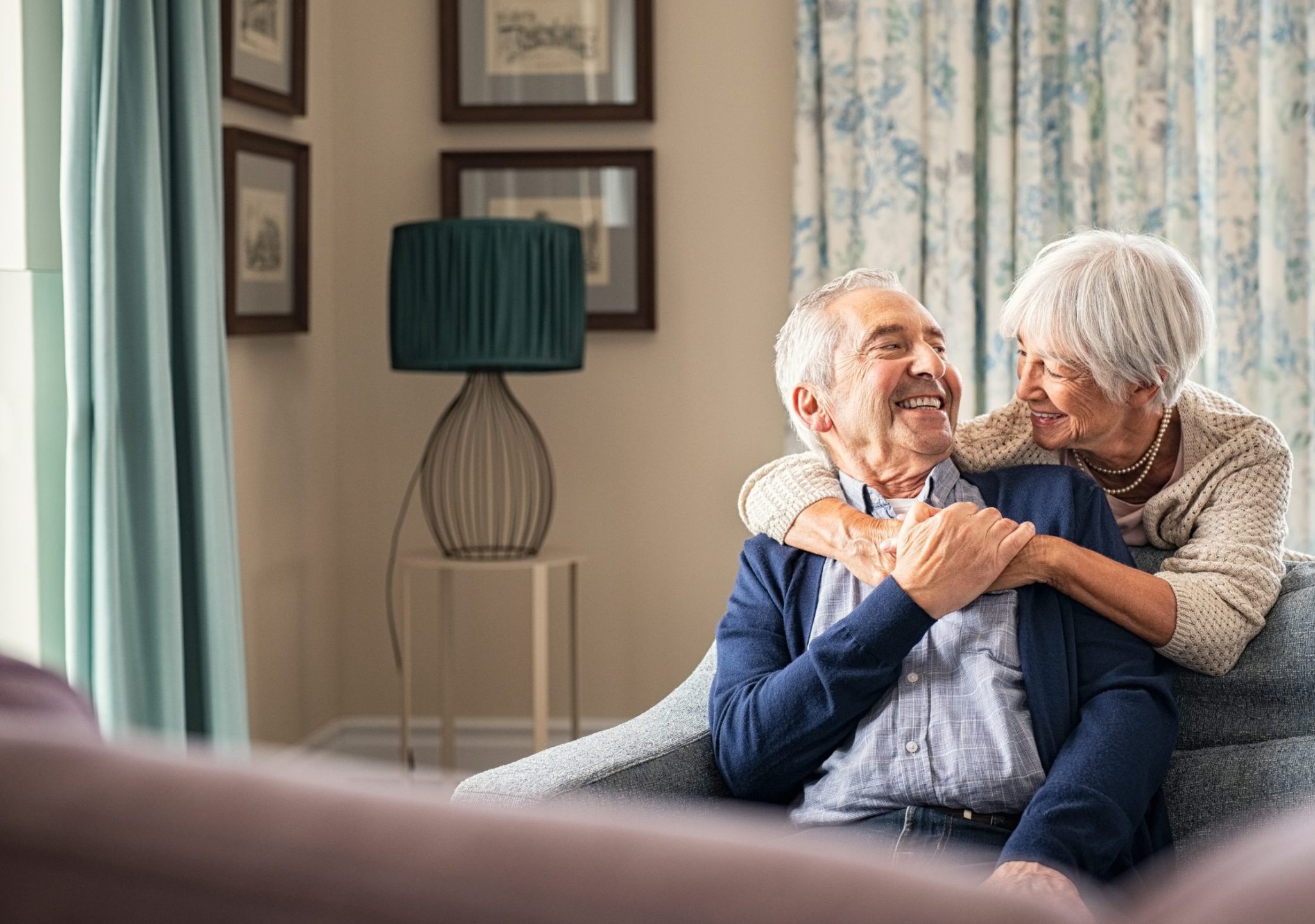 An elderly woman hugs a smiling elderly man from behind as they sit on a couch in a cozy, sunlit living room—an inviting space ideal for pre-memory care, decorated with floral curtains and framed pictures.