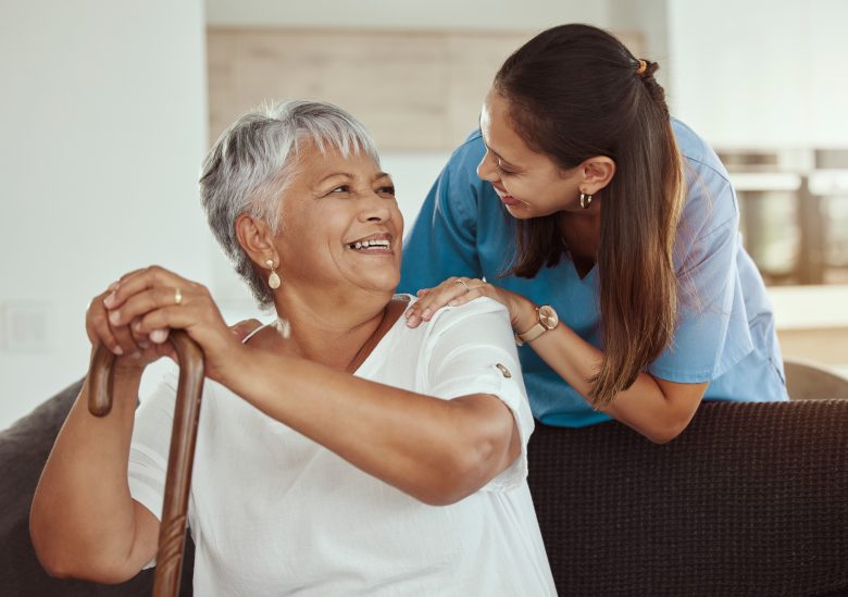 An elderly woman with gray hair sits smiling on a couch, holding a cane. A young caregiver in blue scrubs gently rests a hand on her shoulder. Their warm smiles reflect the caring Eskaton Gold River lifestyle they share every day.