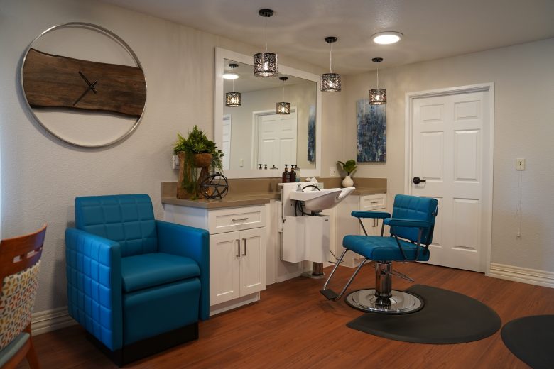 A modern salon interior reflecting the Eskaton Gold River lifestyle, featuring teal chairs, a hair-washing sink, white cabinets, a large mirror, pendant lights, and a wooden wall clock. The wood floor and tidy space create a clean, inviting atmosphere.