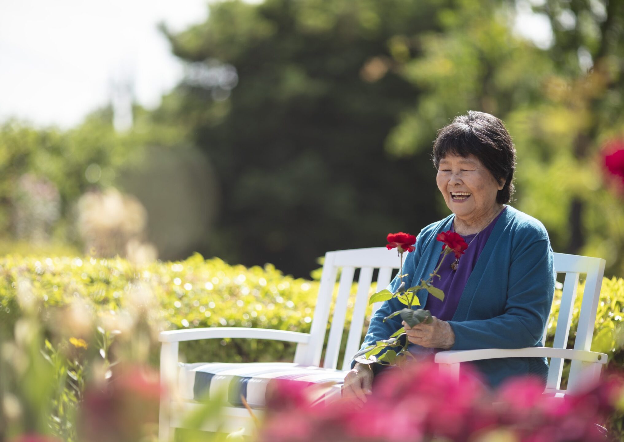 An elderly woman sits on a white bench outdoors at Eskaton Gold River memory care, smiling and holding a red rose, surrounded by greenery and blooming flowers on a sunny day.