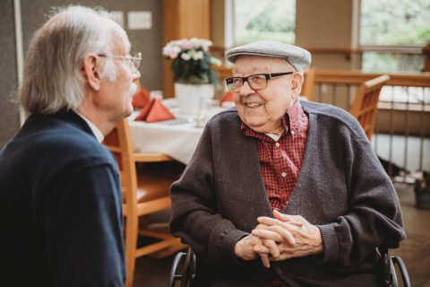 An elderly man in a wheelchair, wearing glasses, a cap, and a cardigan, smiles warmly while chatting with another older man in the bright, cozy Eskaton Gold River senior living dining room adorned with flowers.