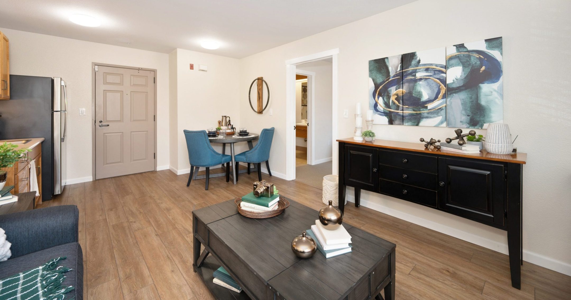 A modern, bright living room with wood flooring, a coffee table, black sideboard, abstract wall art, a small dining table with two blue chairs, and a view into the kitchen and hallway.