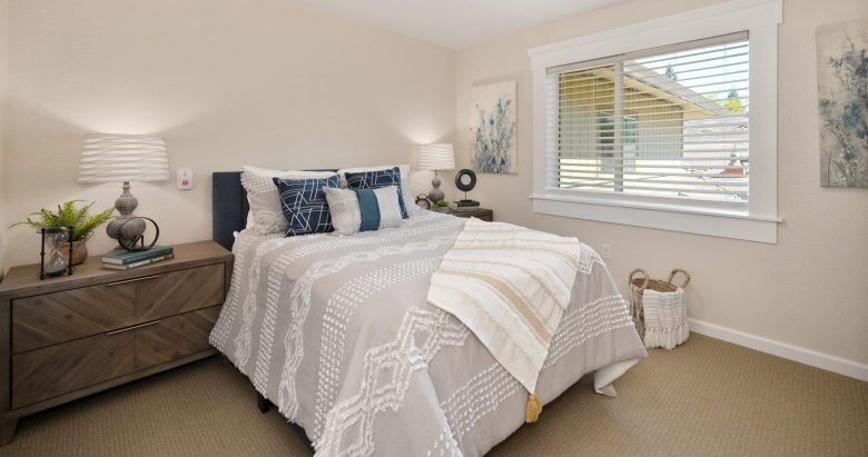 A cozy bedroom with a neatly made bed, decorative pillows, two modern nightstands with lamps, a window with blinds letting in natural light, and a basket in the corner on a beige carpeted floor.