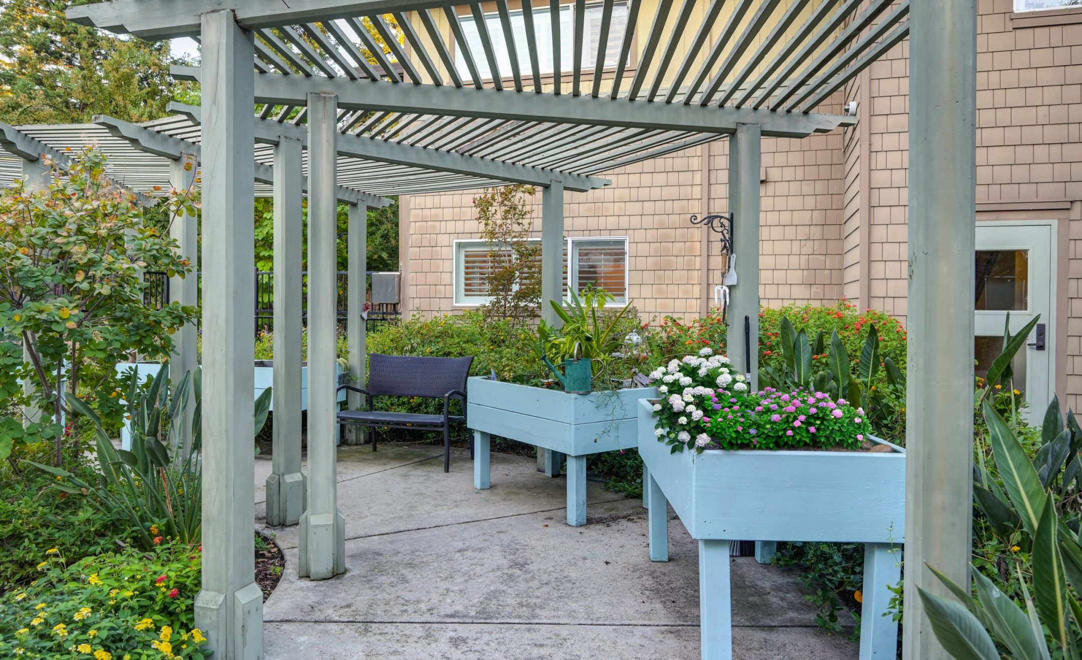 A wooden pergola with raised blue planter boxes filled with flowers and greenery lines a paved walkway next to a tan building—capturing the inviting Eskaton Gold River lifestyle. A black bench sits in the background.