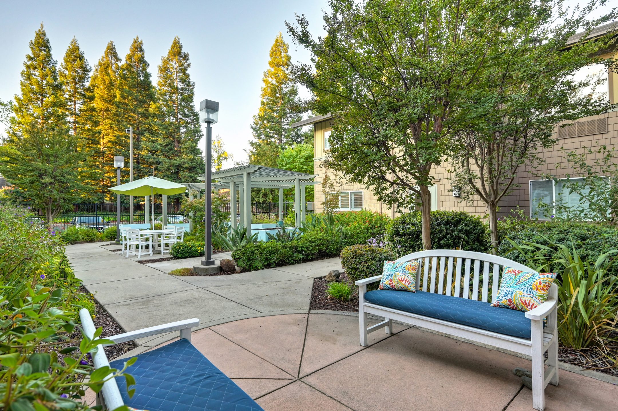 Experience the Eskaton Gold River lifestyle in a peaceful outdoor garden with white benches, blue cushions, colorful pillows, a gazebo with a green umbrella, lush trees and shrubs, and a paved walkway near a beige building.