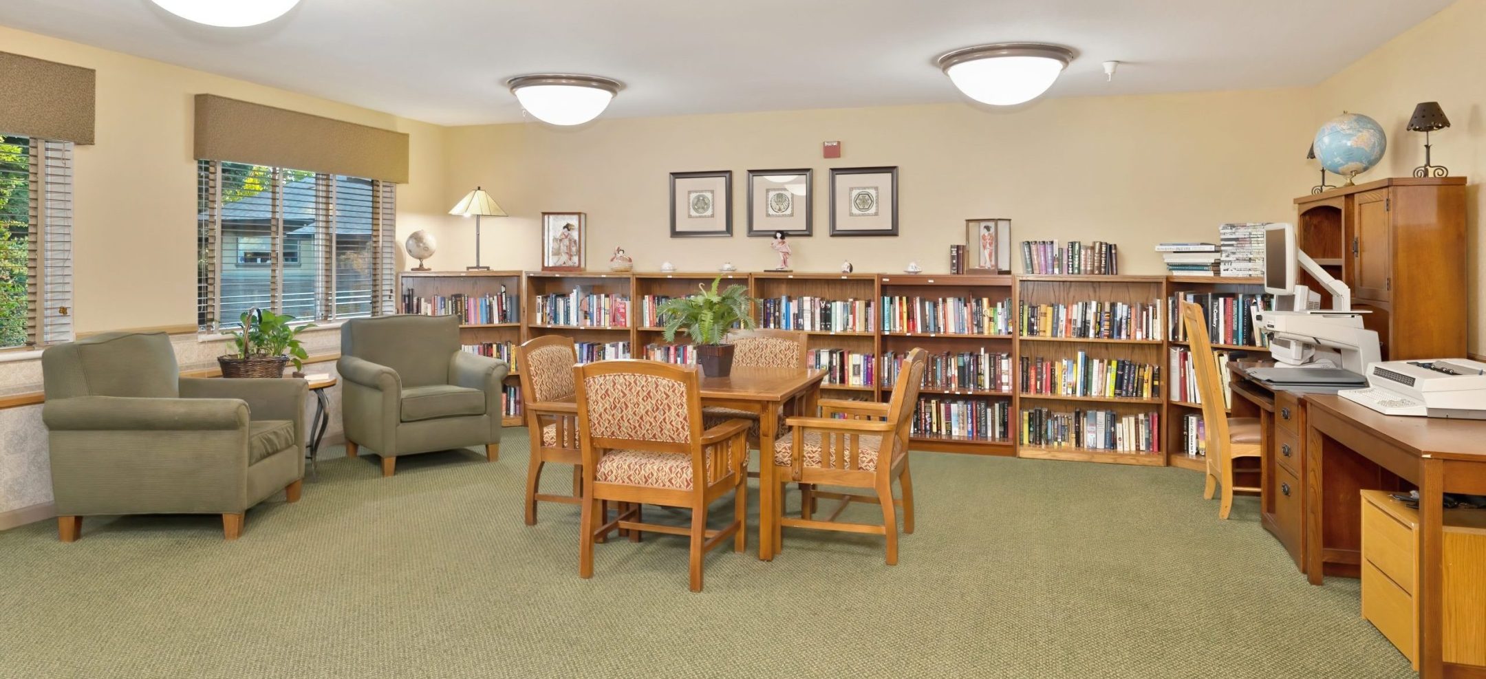 A cozy library room at Eskaton Gold River memory care features bookshelves, a central table with chairs, armchairs by the windows, potted plants, framed pictures on the walls, and a desk with office equipment on the right side.