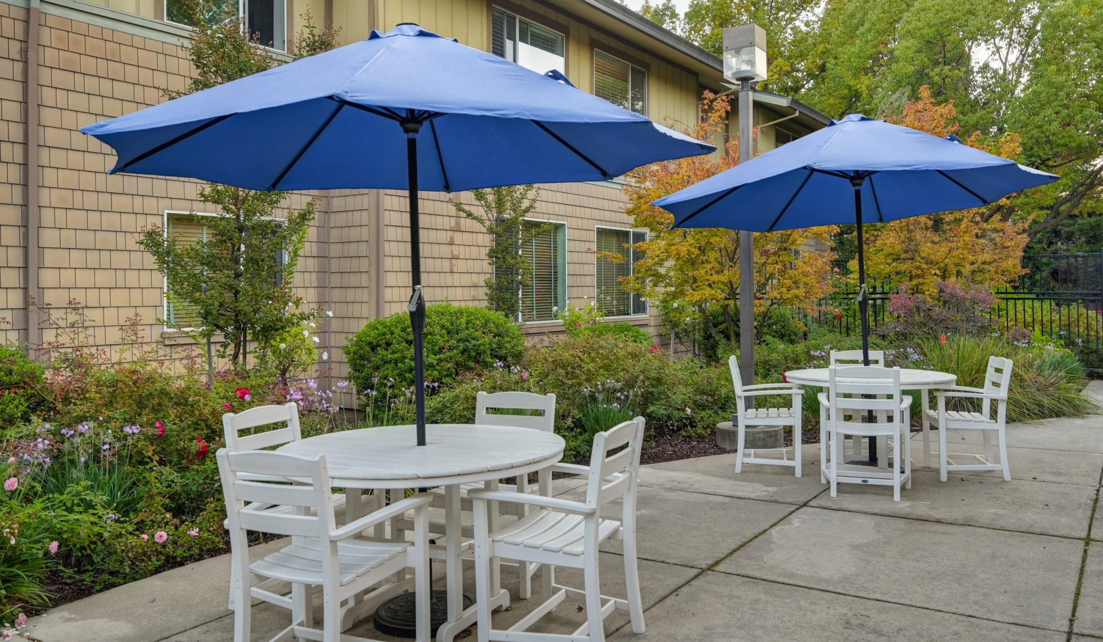 Outdoor patio with white tables and chairs, each shaded by a large blue umbrella. Surrounded by greenery, flowers, and trees next to a beige building with windows, it’s an inviting spot for Eskaton Gold River dining.