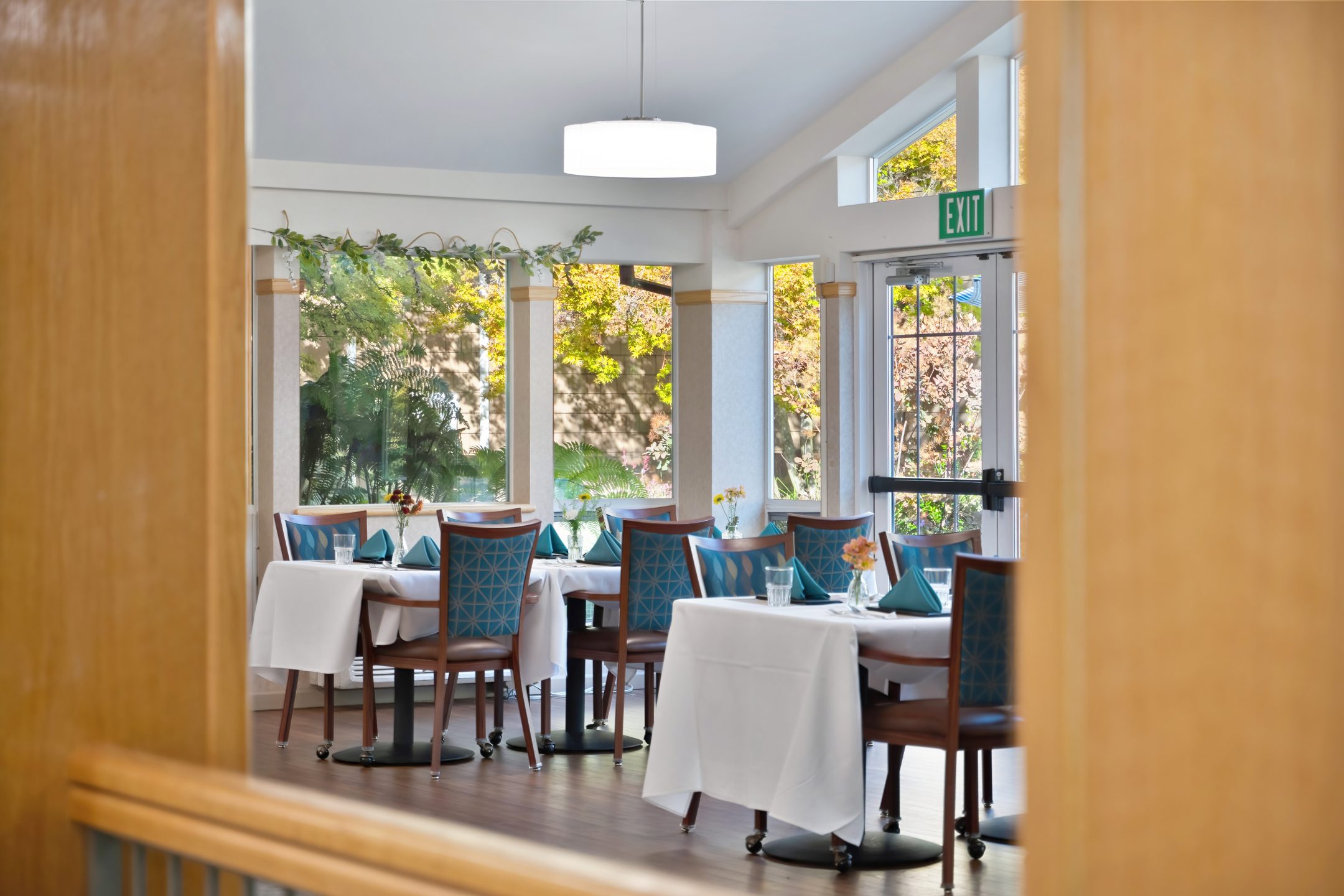 A bright, elegant dining room reflecting the Eskaton Gold River lifestyle, with tables covered in white cloths, teal napkins and flowers, blue patterned chairs, large windows, and sunlight streaming past an EXIT sign above a glass door.