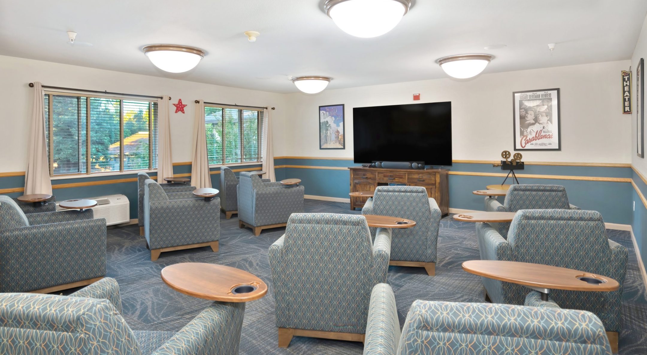 A spacious media room reflects the Eskaton Gold River lifestyle, with patterned armchairs facing a large flat-screen TV on a wooden stand. Small tables are attached to each chair, natural light streams through windows, and movie posters adorn the walls.