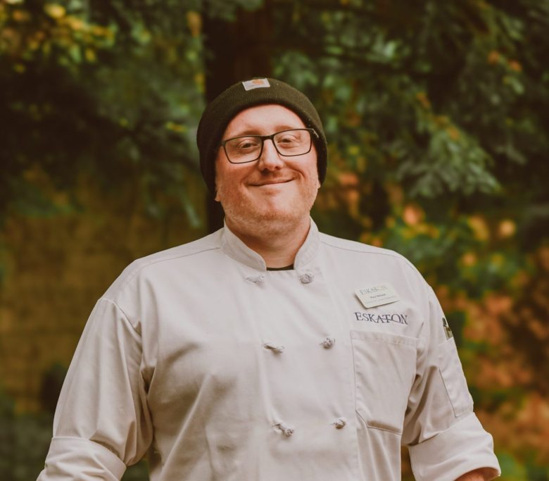 A smiling man wearing glasses, a beanie, and a chef's jacket stands outdoors with greenery in the background. Showcasing his tattooed arm, he embodies the passion behind Eskaton Gold River dining.