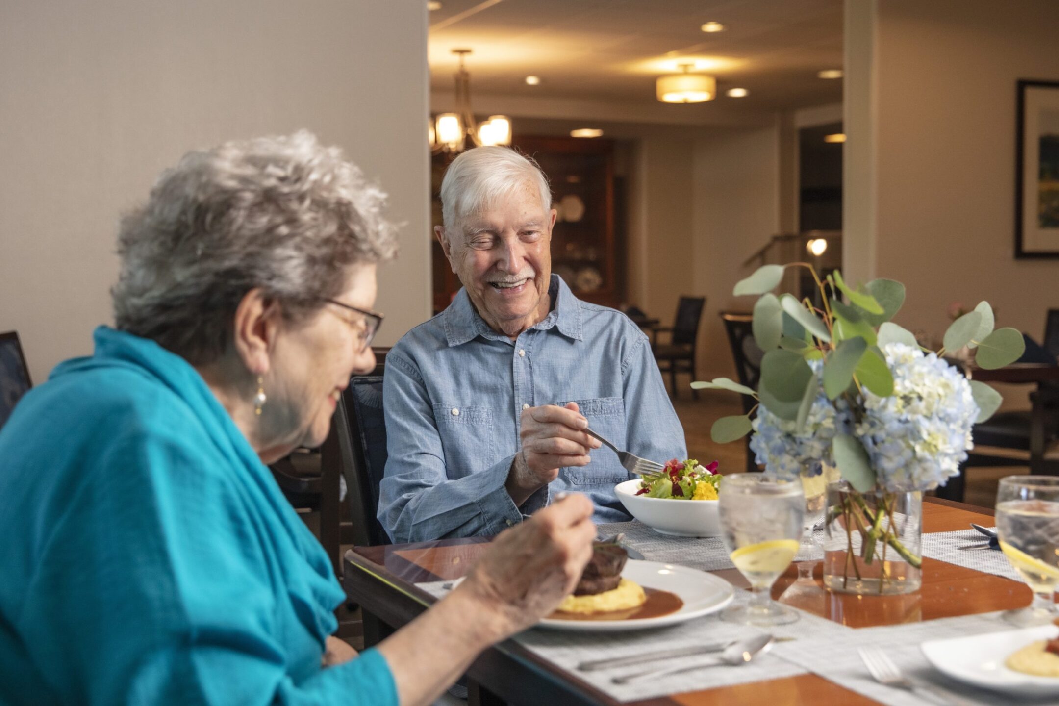 Two older adults sit at a dining table, smiling and enjoying a meal together in a warmly lit restaurant during a Lunch and Learn. A vase of flowers and glasses of water with lemon slices add to the inviting scene.