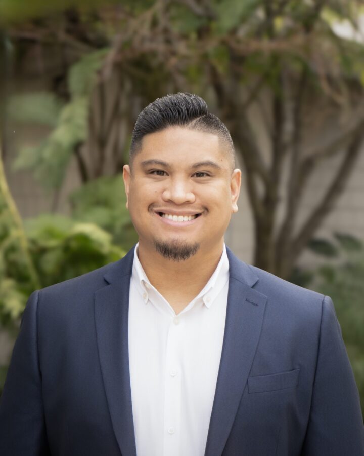 A man with short dark hair and a neatly trimmed beard smiles while wearing a navy blazer and white shirt, standing outdoors amid the lush greenery of Eskaton Gold River.