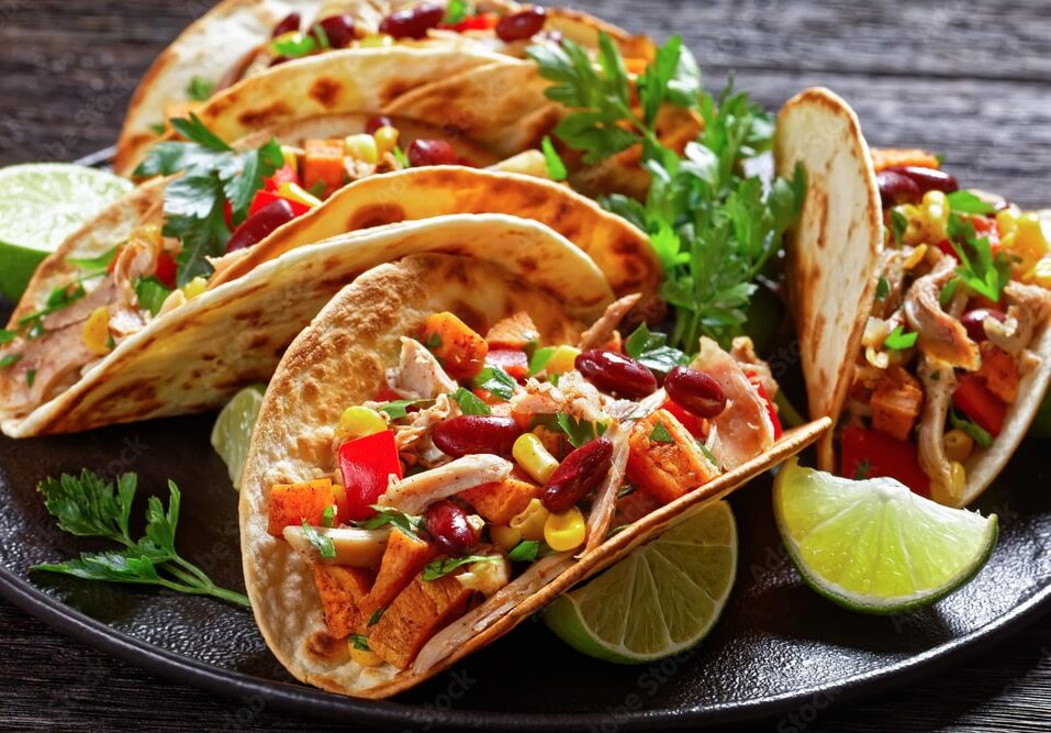 A plate of grilled tacos perfect for the Big Day of Tacos, filled with shredded meat, kidney beans, corn, diced red peppers, and herbs, garnished with fresh parsley and lime wedges on a dark wooden table.