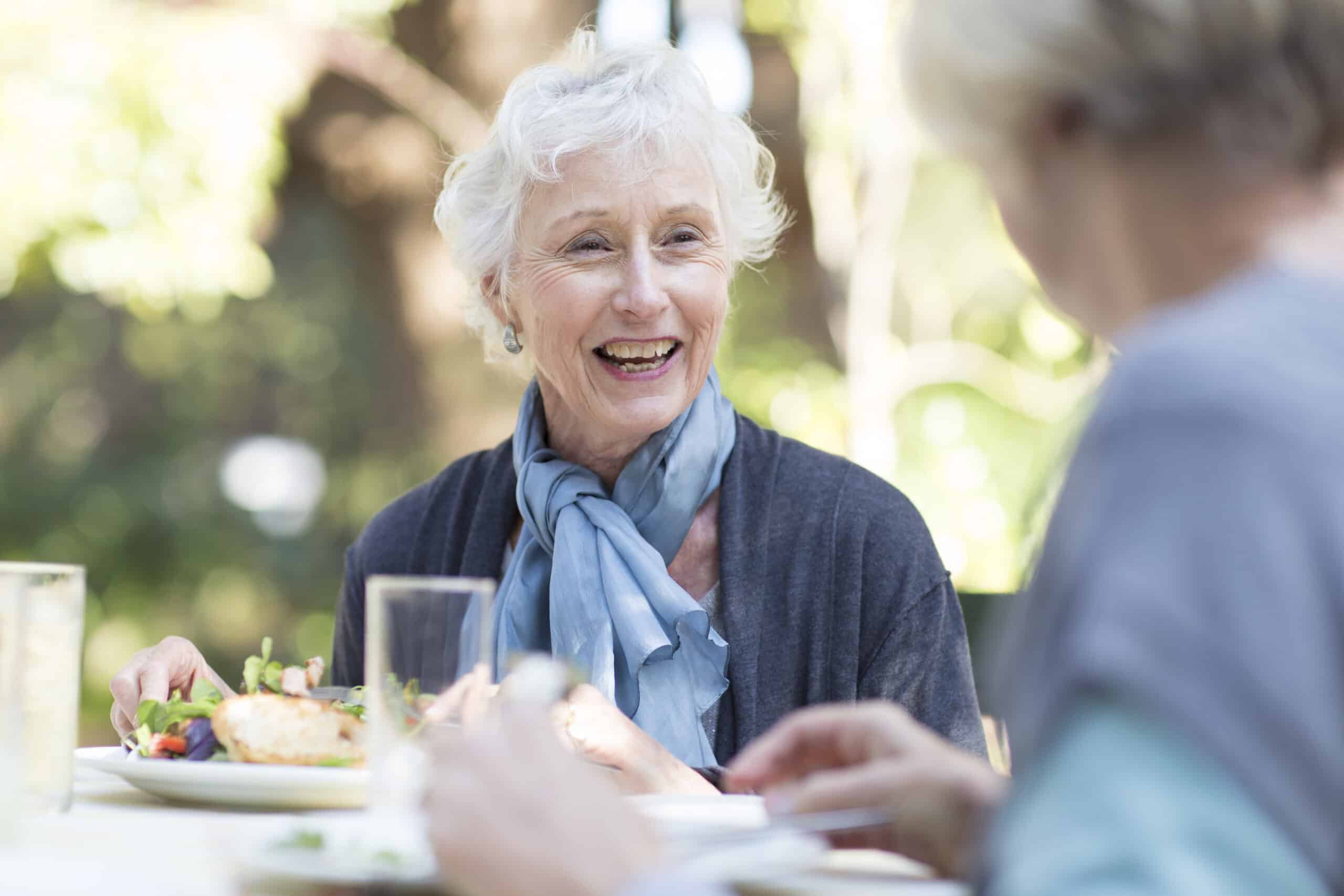 An older woman with short white hair and a blue scarf smiles while dining at an outdoor table, enjoying a meal with another person. The background is blurred with greenery, creating a warm scene of shared discovery.