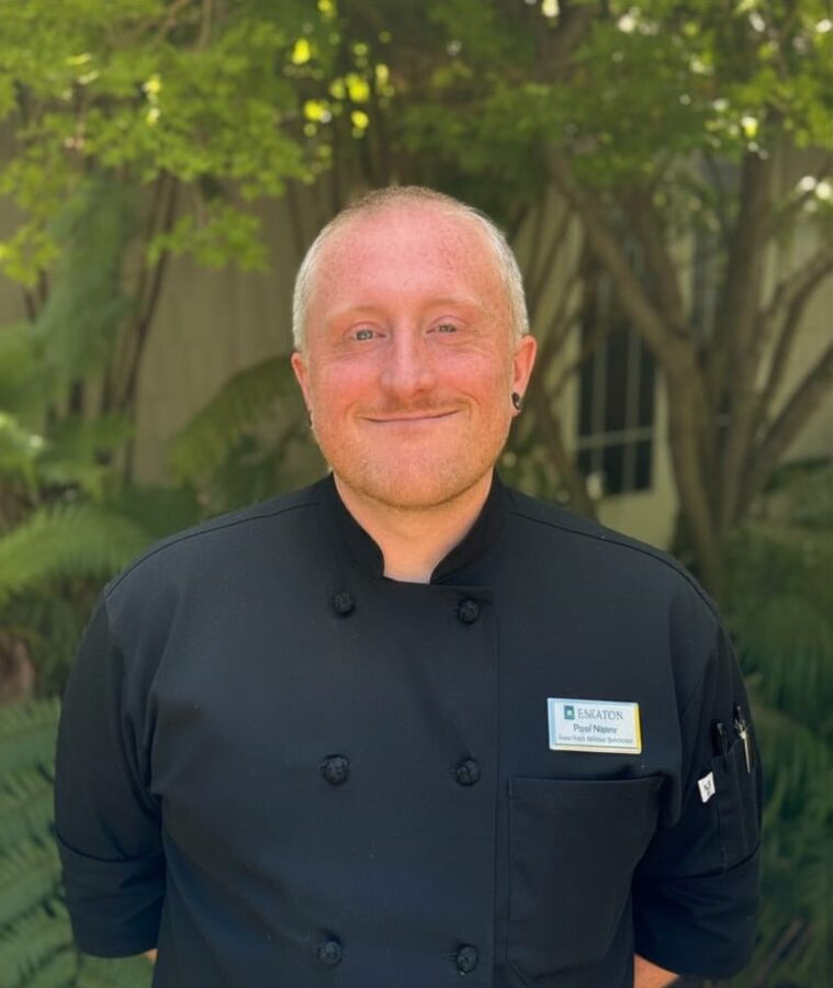 A smiling chef with short light hair, wearing a black chef’s coat and name tag, stands outdoors in front of green foliage and trees on a sunny day, ready to share his culinary experience.