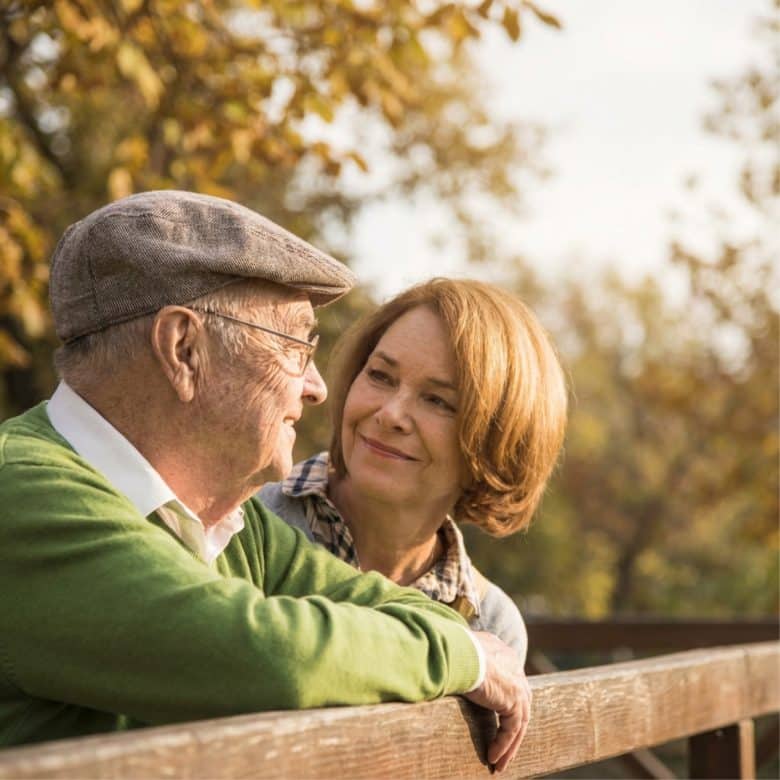 An elderly man in a flat cap and a woman with short hair smile at each other while leaning on a wooden fence outdoors on a sunny day, enjoying the vibrant Eskaton Gold River lifestyle amid autumn trees in the background.