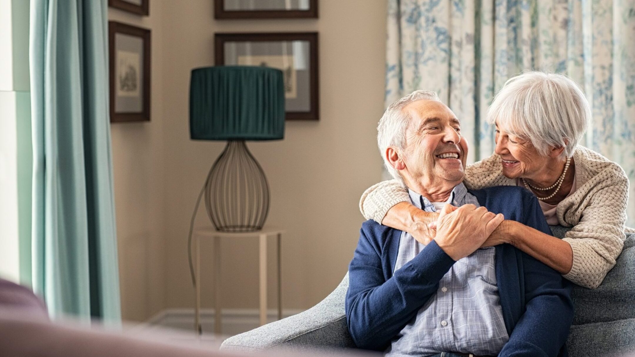Join Eskaton Granite Bay’s Parkinson’s Support Group for connection. An elderly couple sits on a couch at home, smiling and embracing warmly, with the woman hugging the man from behind. The room is softly lit with patterned curtains and framed pictures on the wall.