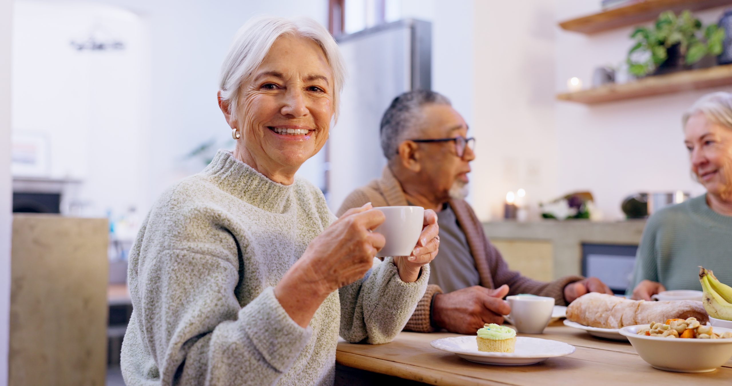 An older woman with gray hair smiles and holds a coffee cup at a table, enjoying food and drinks with two elderly friends in a cozy, well-lit kitchen—perfect for Short-Term Stays filled with warmth and connection.