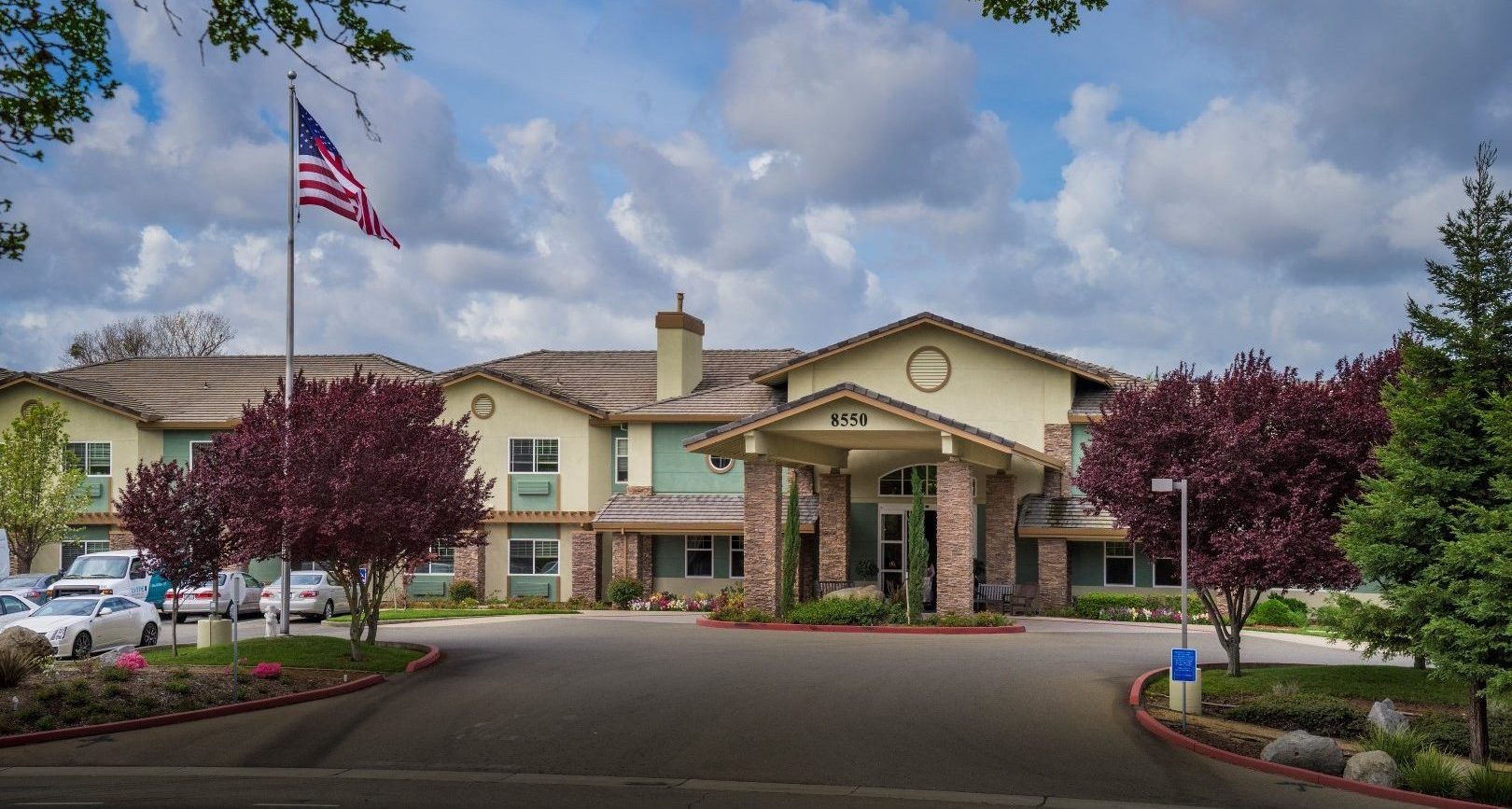 A large Independent Living building with stone columns and a covered entrance is surrounded by trees and landscaping under a partly cloudy sky. An American flag flies on a flagpole in front, and the address 8650 is visible above the entrance.