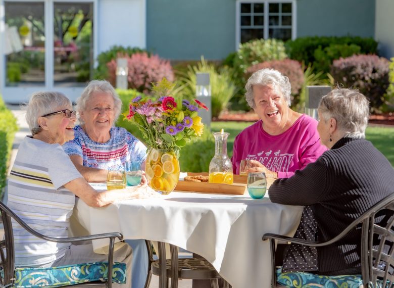 Four smiling elderly women sit around a table outdoors, enjoying drinks and conversation. A pitcher of lemon water and a vase of flowers are on the table. Greenery and a building are visible in the background.