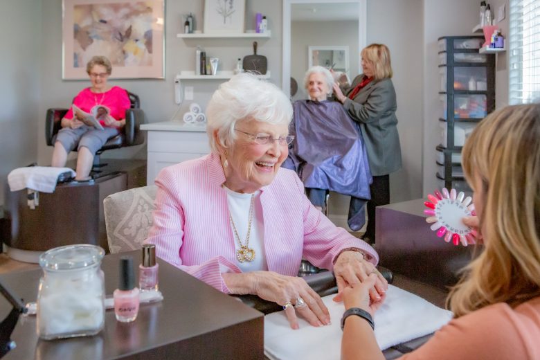 Two elderly women are at a salon; one gets her nails done and smiles, while another has her hair styled. A stylist and another woman are assisting. The room is bright and welcoming, with beauty products on the counter.