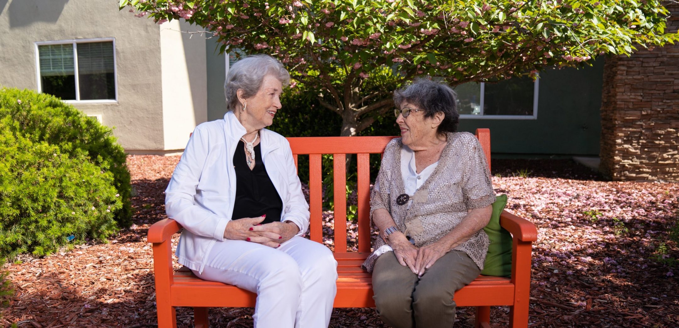 Two elderly women sit and talk on an orange bench in a garden, surrounded by mulch and greenery, discussing various living options as sunlight filters through the trees. They appear relaxed and engaged in conversation. Contact Eskaton Granite Bay
