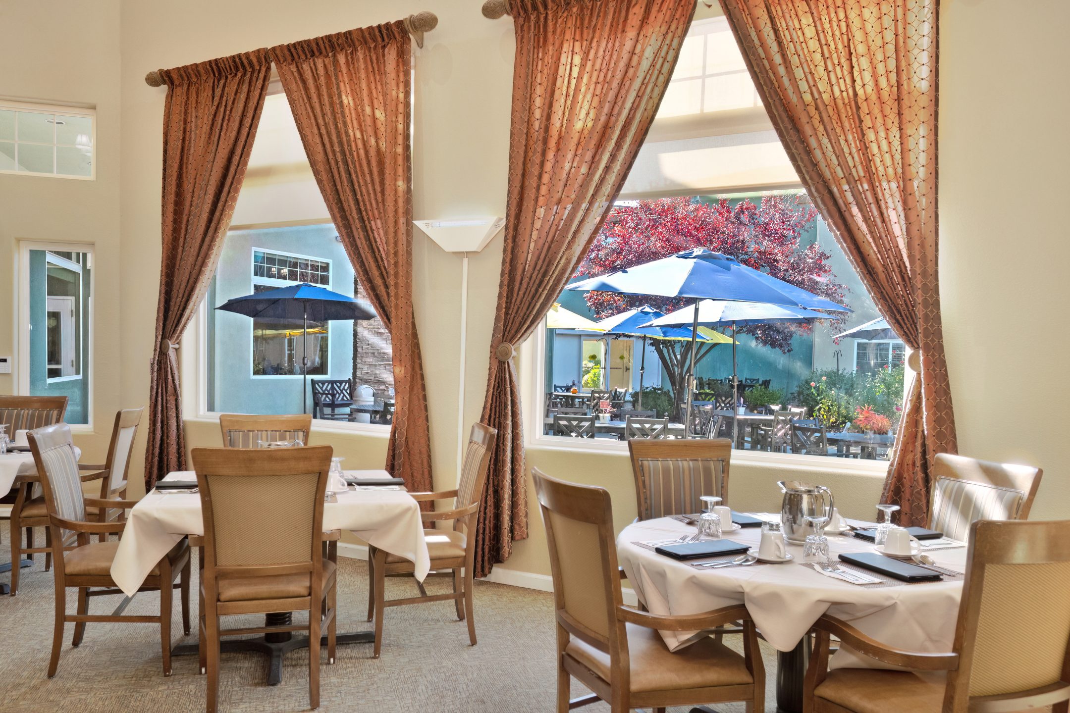 A dining room with round tables set for meals, wooden chairs, and large windows with brown curtains. Outside, there is a patio with umbrellas, tables, chairs, and a red-leaved tree. Natural light fills the room.