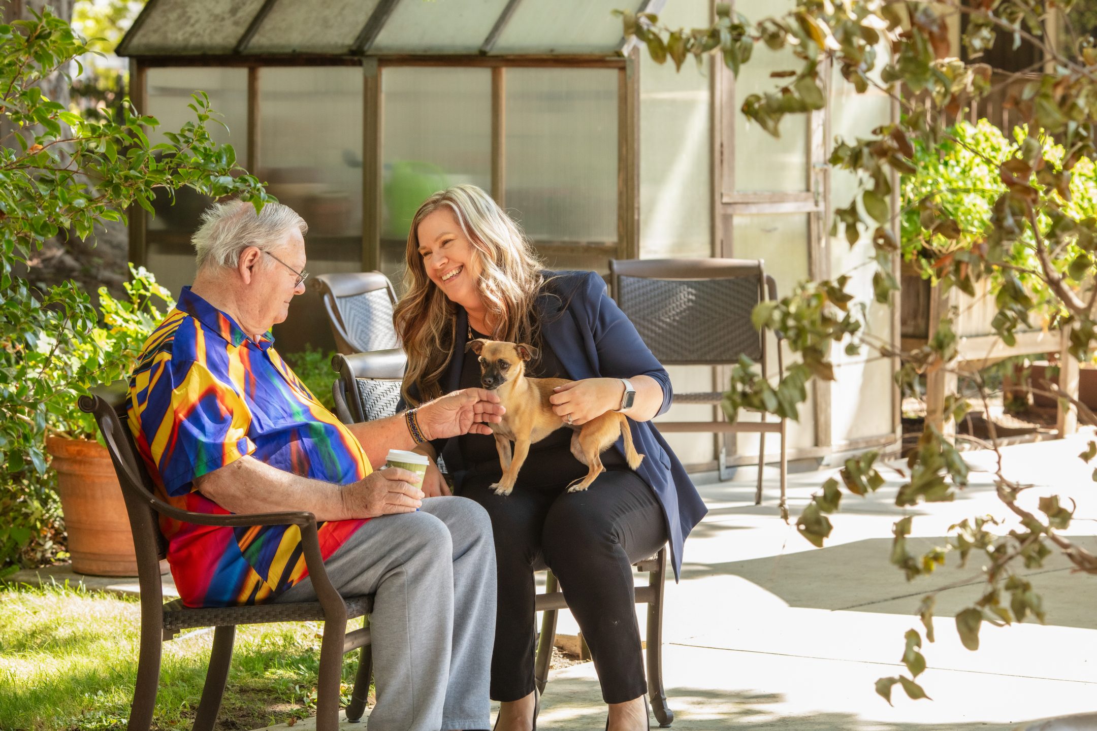 An older man and a woman sit outdoors on a sunny day at Eskaton Granite Bay assisted living, smiling and petting a small dog. The man holds a cup as they relax near a greenhouse surrounded by greenery.