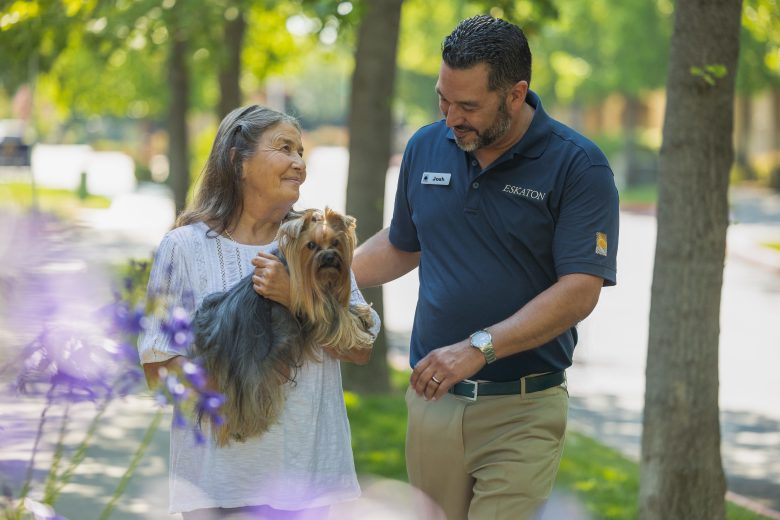 An elderly woman holding a small dog walks outside with a smiling man in a navy polo shirt and name tag. At Eskaton Granite Bay senior living, they are surrounded by green trees and sunlight, enjoying a pleasant day together.