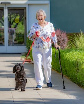 An elderly woman smiles as she walks outside at Eskaton Granite Bay senior living, leading a black dog on a leash and using a cane. She wears a floral top and white pants, with green hedges and a building behind her.