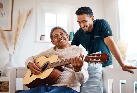 An older man sits on a bench playing an acoustic guitar and smiling, while a younger man stands beside him, leaning on the bench and smiling warmly in the bright, cozy room at Eskaton Granite Bay senior living.