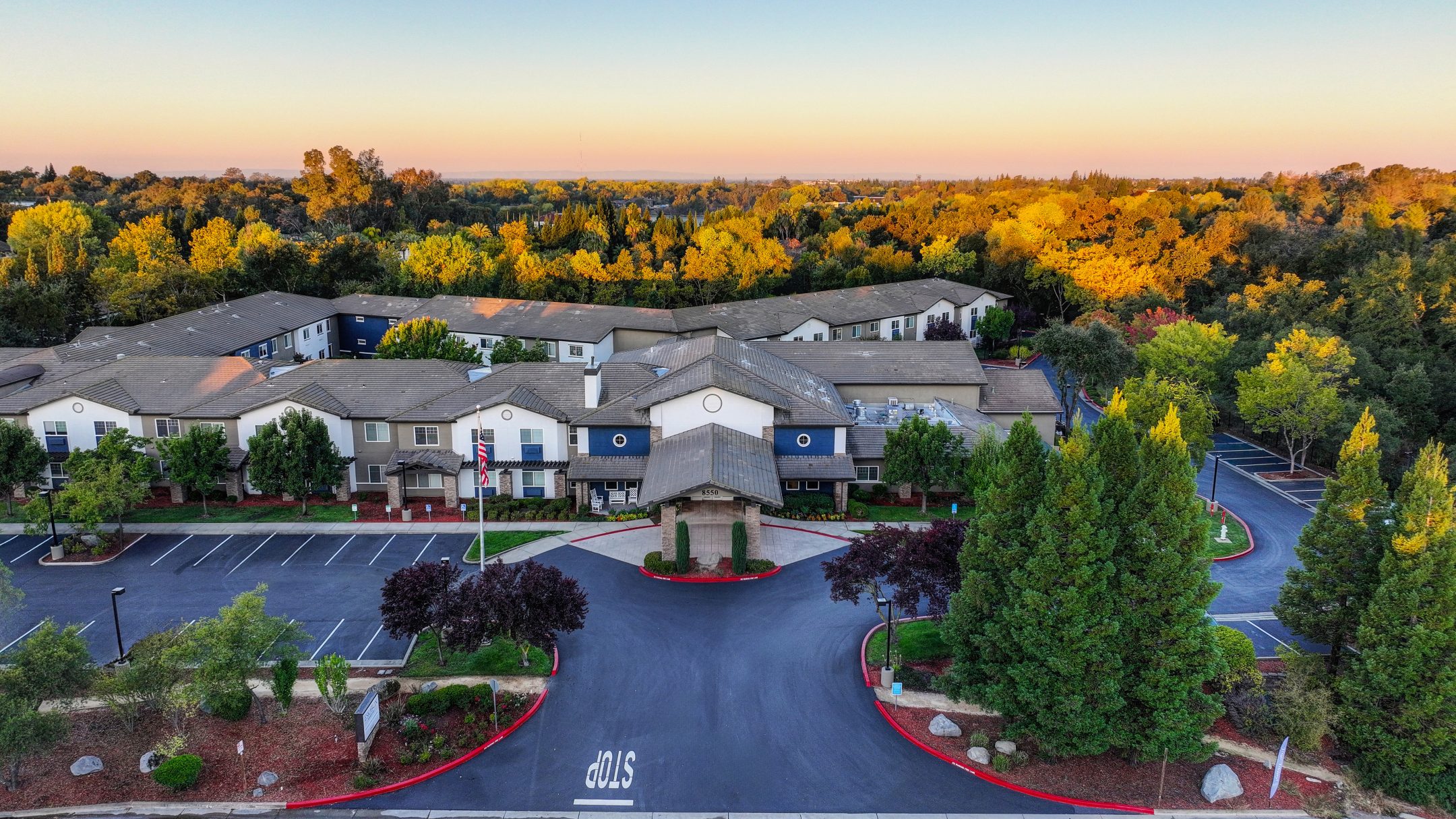 Aerial view of Eskaton Granite Bay senior living complex surrounded by trees, featuring landscaped gardens, empty parking lots, and a main entrance facing a circular driveway under a clear sky at sunset.