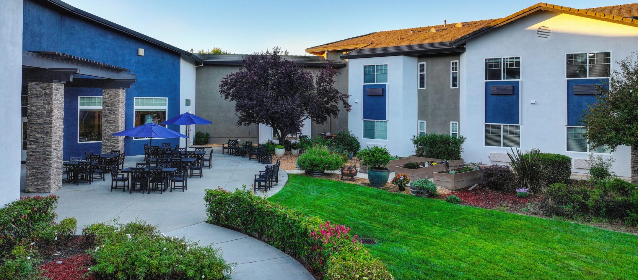 A courtyard at Eskaton Granite Bay senior living features patio tables, blue umbrellas, and chairs nestled between modern buildings, surrounded by green grass, lush plants, and trees under a clear blue sky.