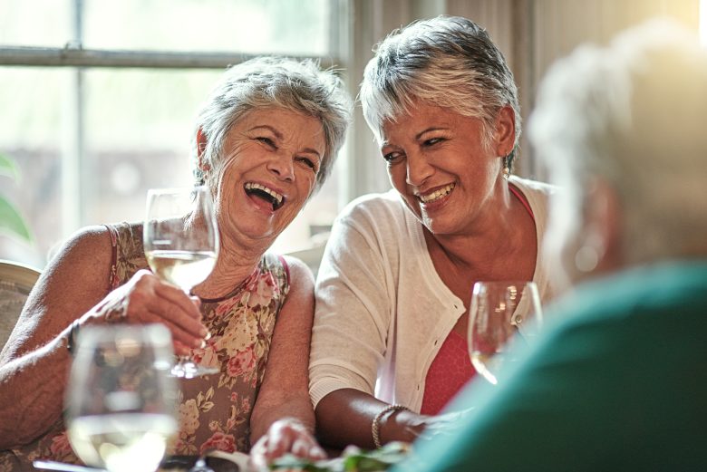 Two older women sit at a table, smiling and laughing with wine glasses in hand, enjoying a meal together at Eskaton Granite Bay dining. Sunlight streams through the window behind them as they savor each other’s company.