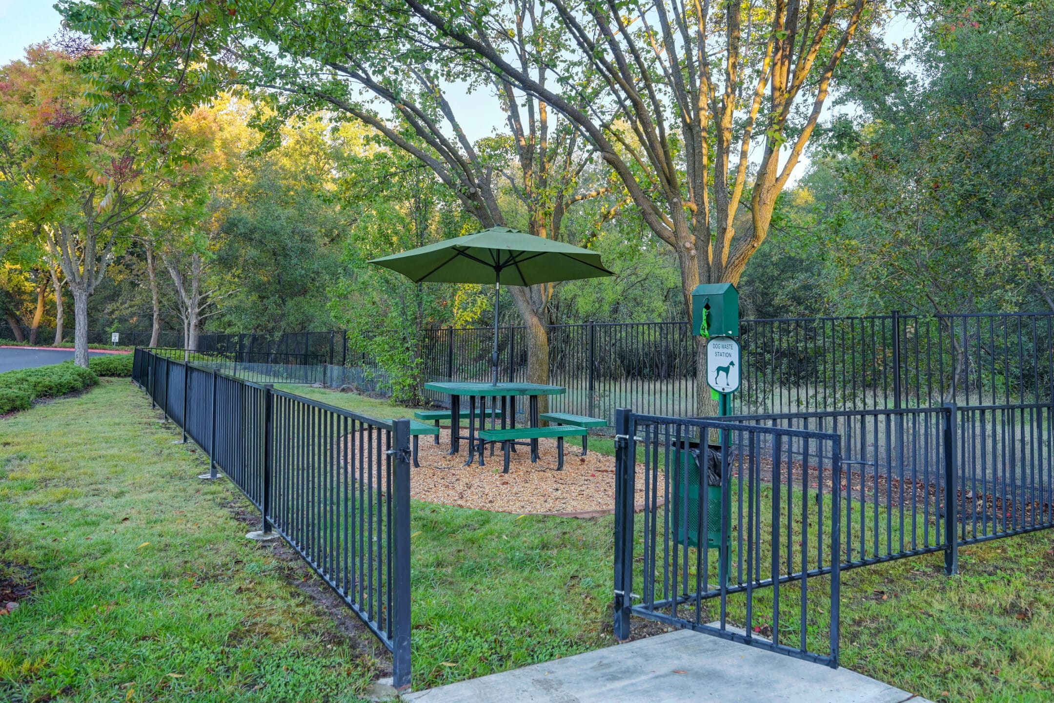 A fenced-in outdoor area at Eskaton Granite Bay senior living features a green picnic table with an umbrella, surrounded by trees. Near the entrance, a dog waste station with a bag dispenser and trash can is provided for residents' convenience.