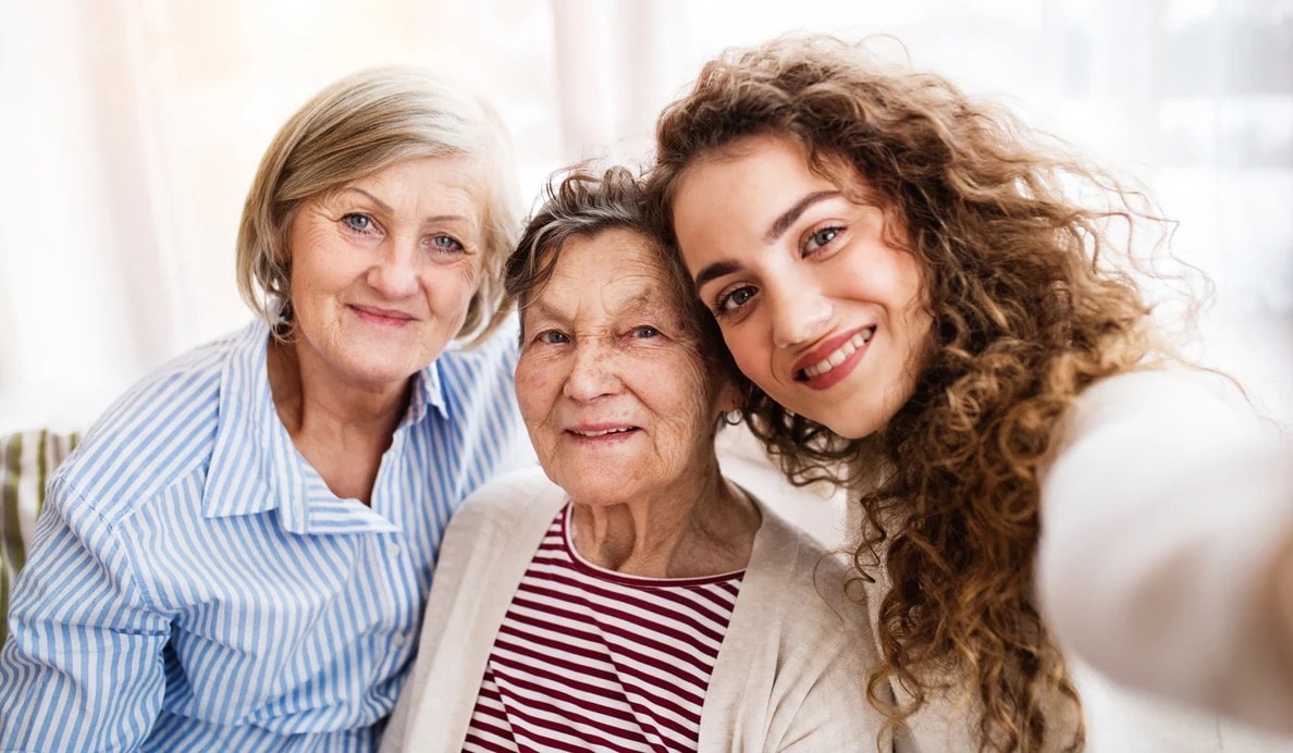 Three women of different generations, likely family members, smile together for a selfie. The older woman is in the center, with a younger and a middle-aged woman on either side, all looking happy and close.