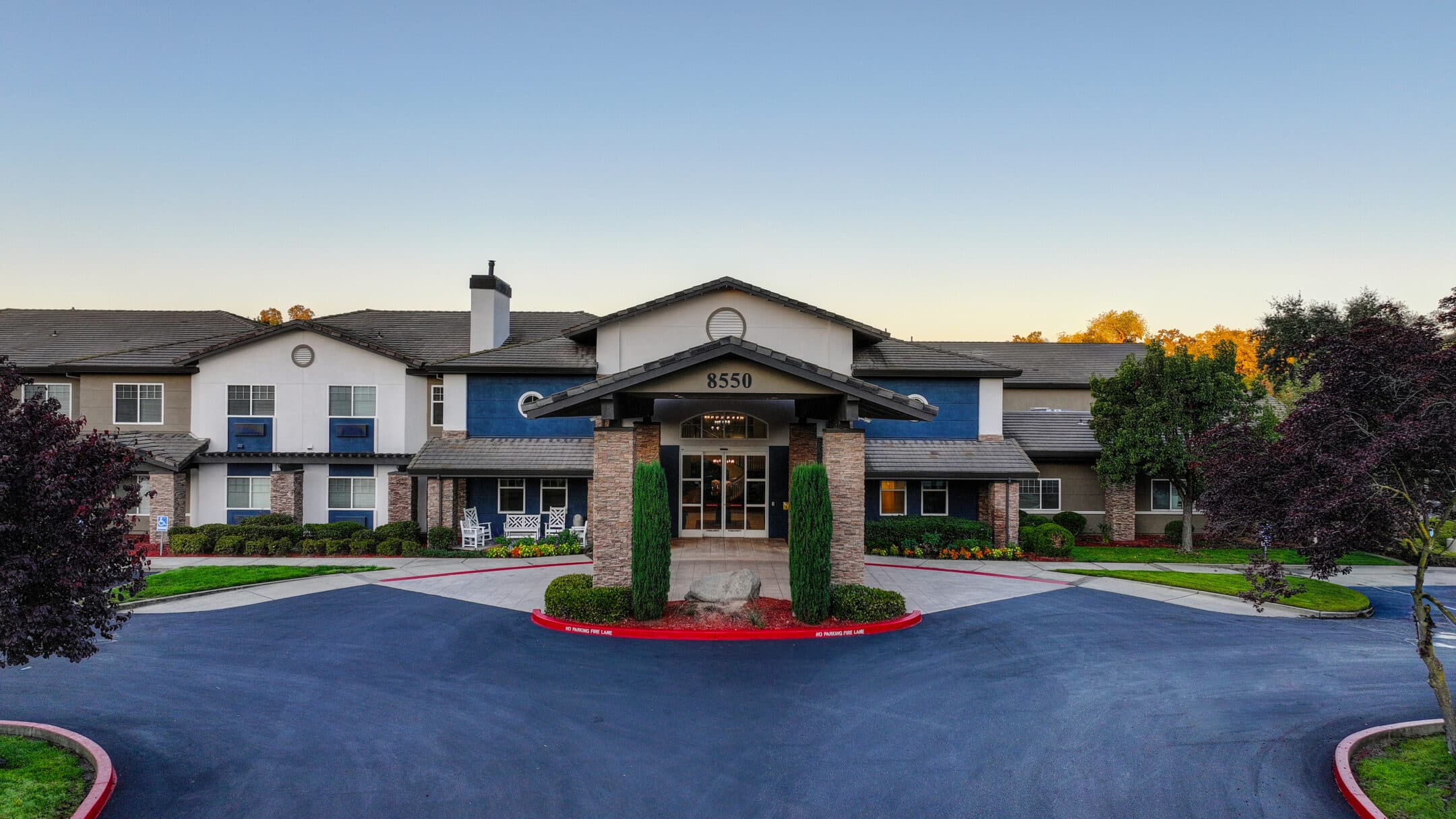 A large, modern building with a covered entrance and columns, surrounded by trimmed bushes, manicured lawns, and trees. The address “8550” is displayed above the entrance beneath a clear sky. Schedule a tour to experience this impressive property firsthand.