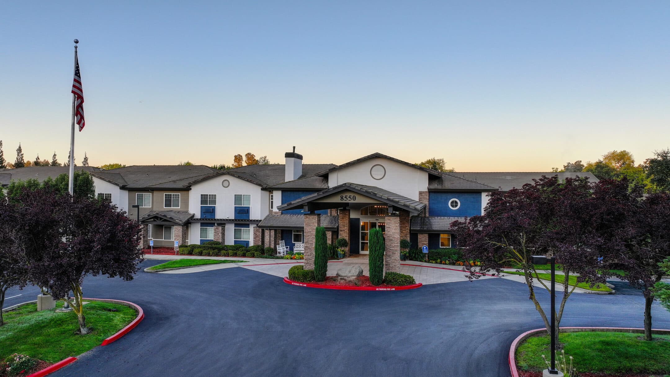 A modern, single-story building with a circular driveway, well-maintained landscaping, and an American flag on a pole out front—captured at sunset under a clear sky. Schedule a Tour to experience this inviting space firsthand.