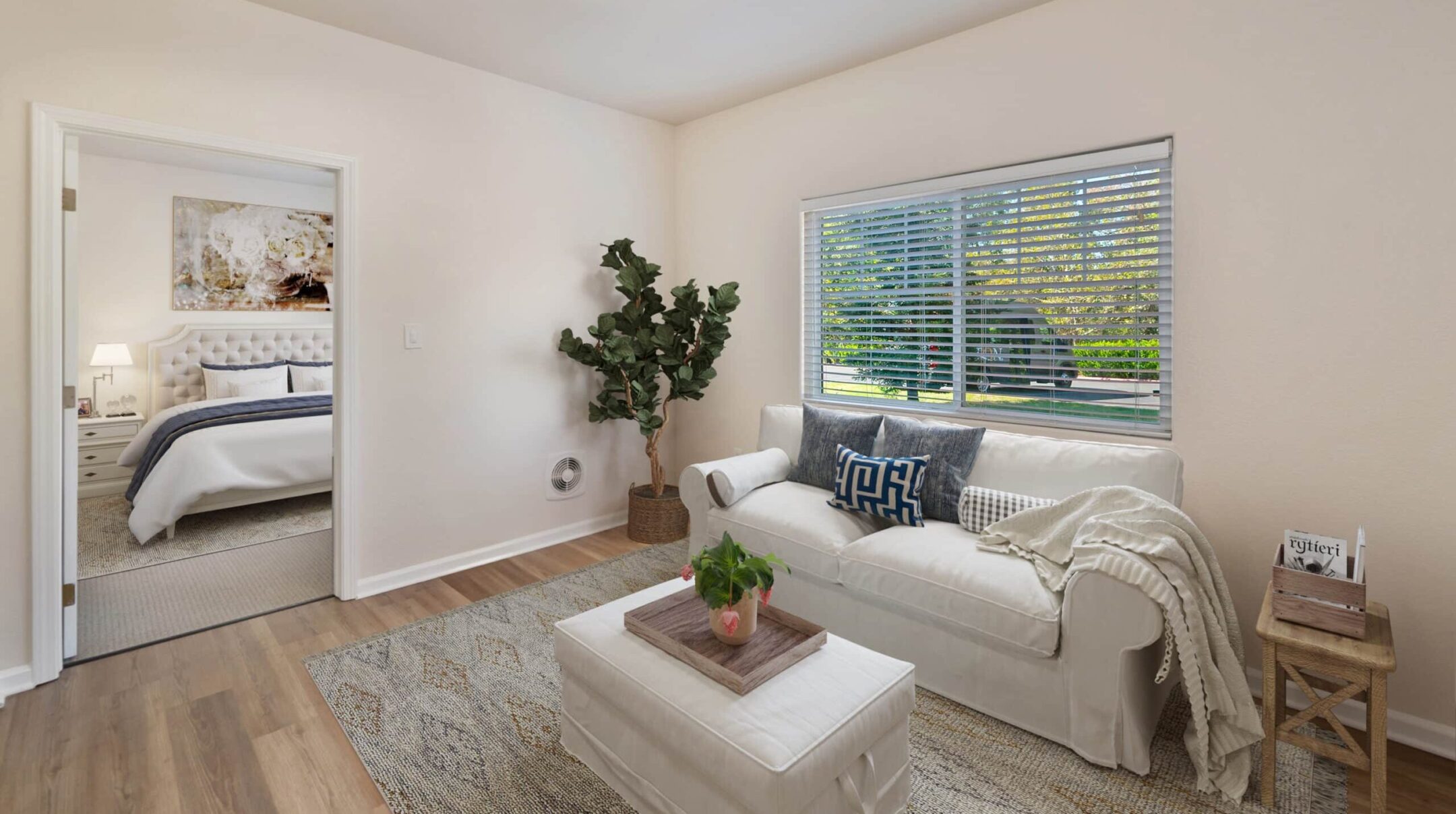 Bright, cozy living room with a white sofa, decorative pillows, potted plant, and a small coffee table. Large window lets in natural light. Bedroom visible through an open door showcases flexible living options. Light walls and wood flooring.