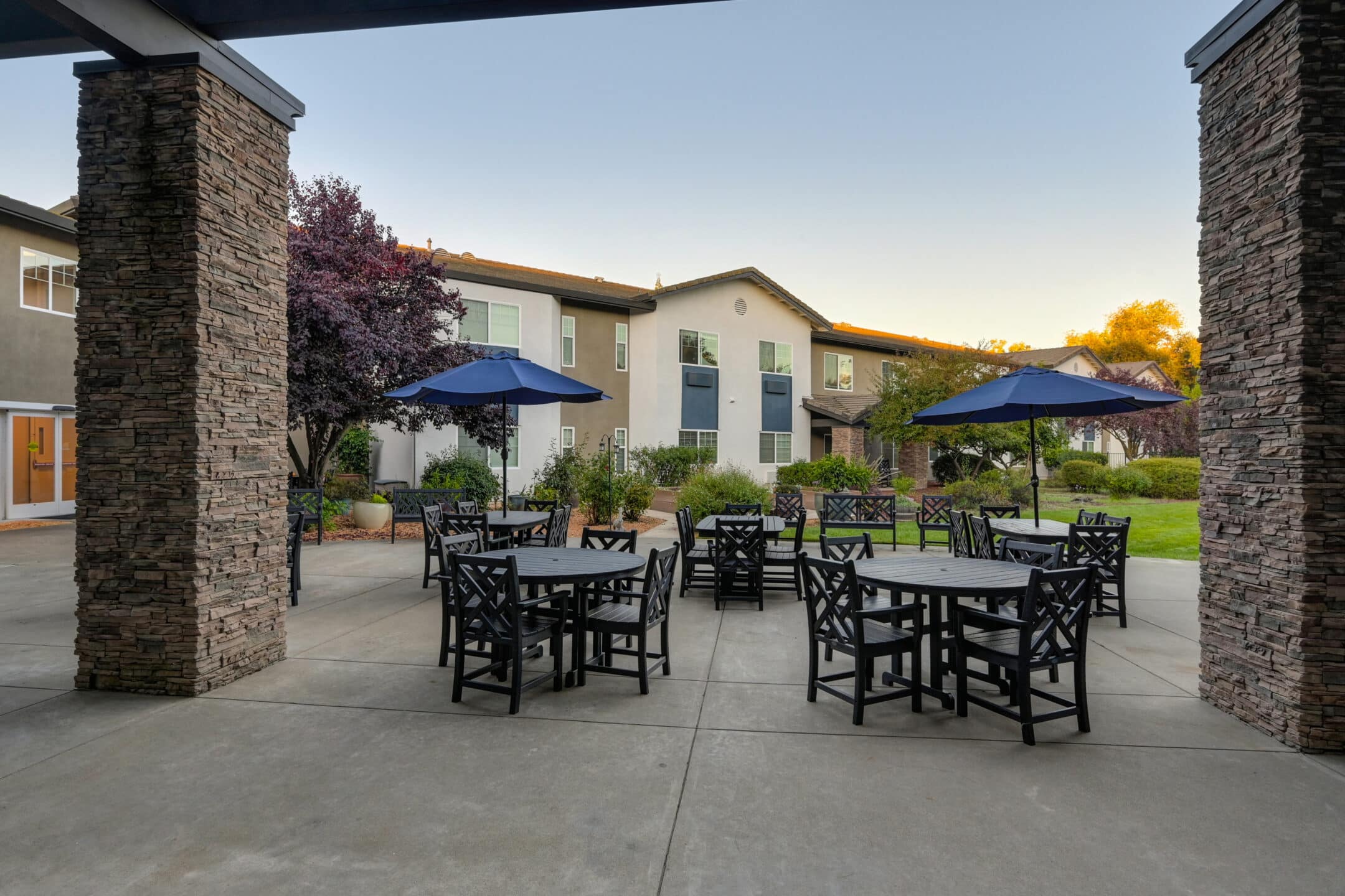 Outdoor patio area with several black tables and chairs, shaded by blue umbrellas, surrounded by stone pillars and facing a landscaped garden at Eskaton Granite Bay senior living, with a two-story building in the background.
