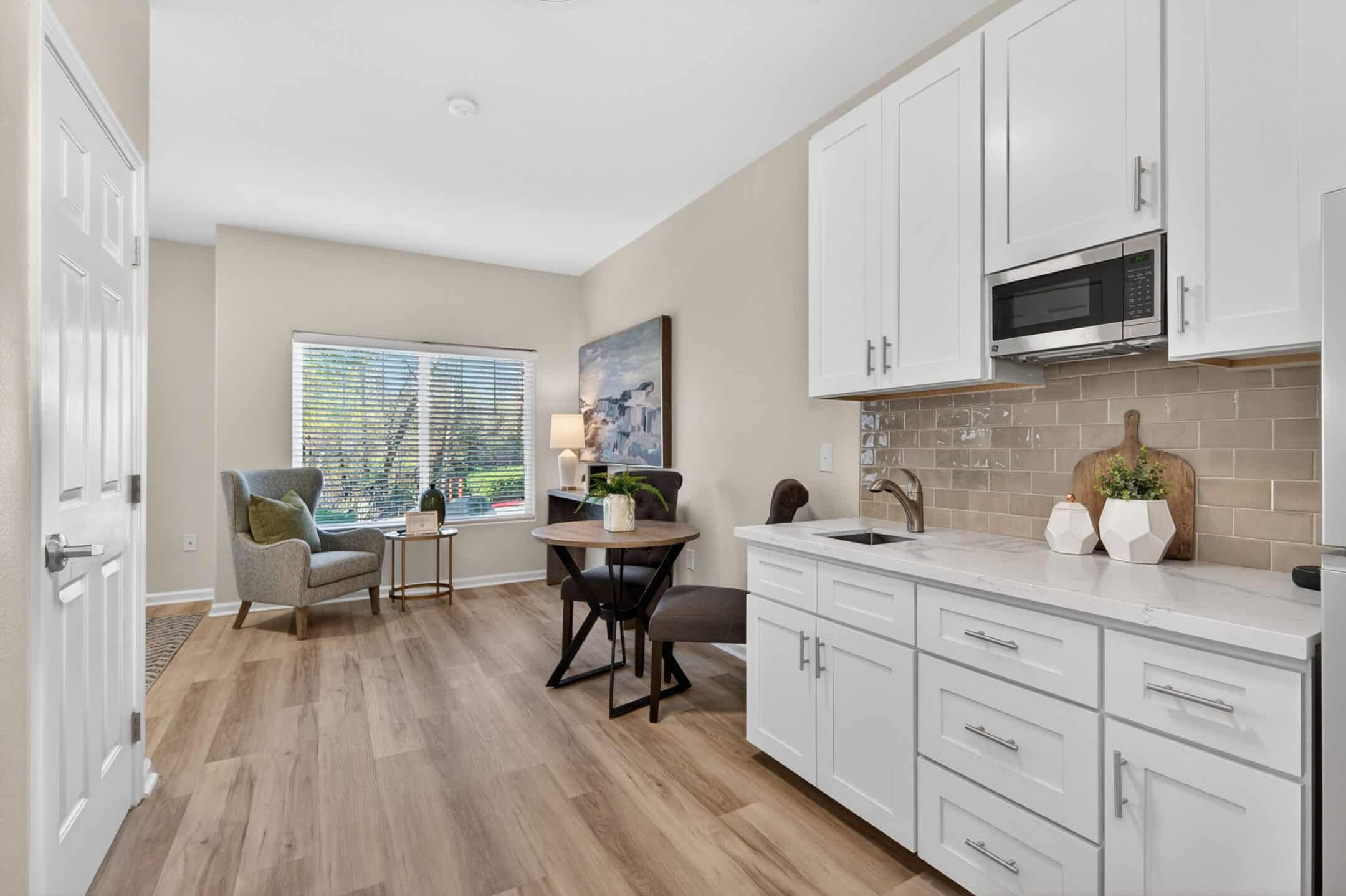 A bright, modern kitchen features white cabinets, a microwave, and light wood floors. A small dining area with a round table and chairs sits near a window with two armchairs and a lamp in the corner.